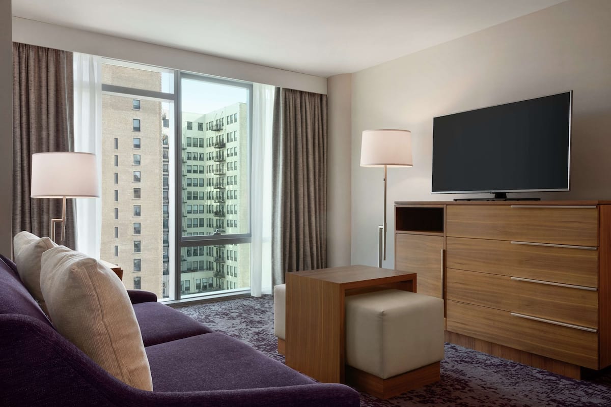 A hotel room with a large window, a sofa, a TV, and a wooden dresser.