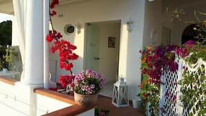 Ein überdachter Außenbereich mit einer weißen Säule, einem Blumentopf mit rosa und weißen Blüten, einem weißen Laternen und einem weißen Gitterzaun mit Blumen.