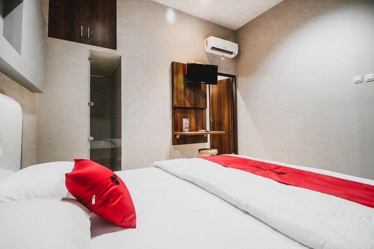 A hotel room with a bed, a red pillow, a wooden cabinet, a shelf, and an air conditioning unit.
