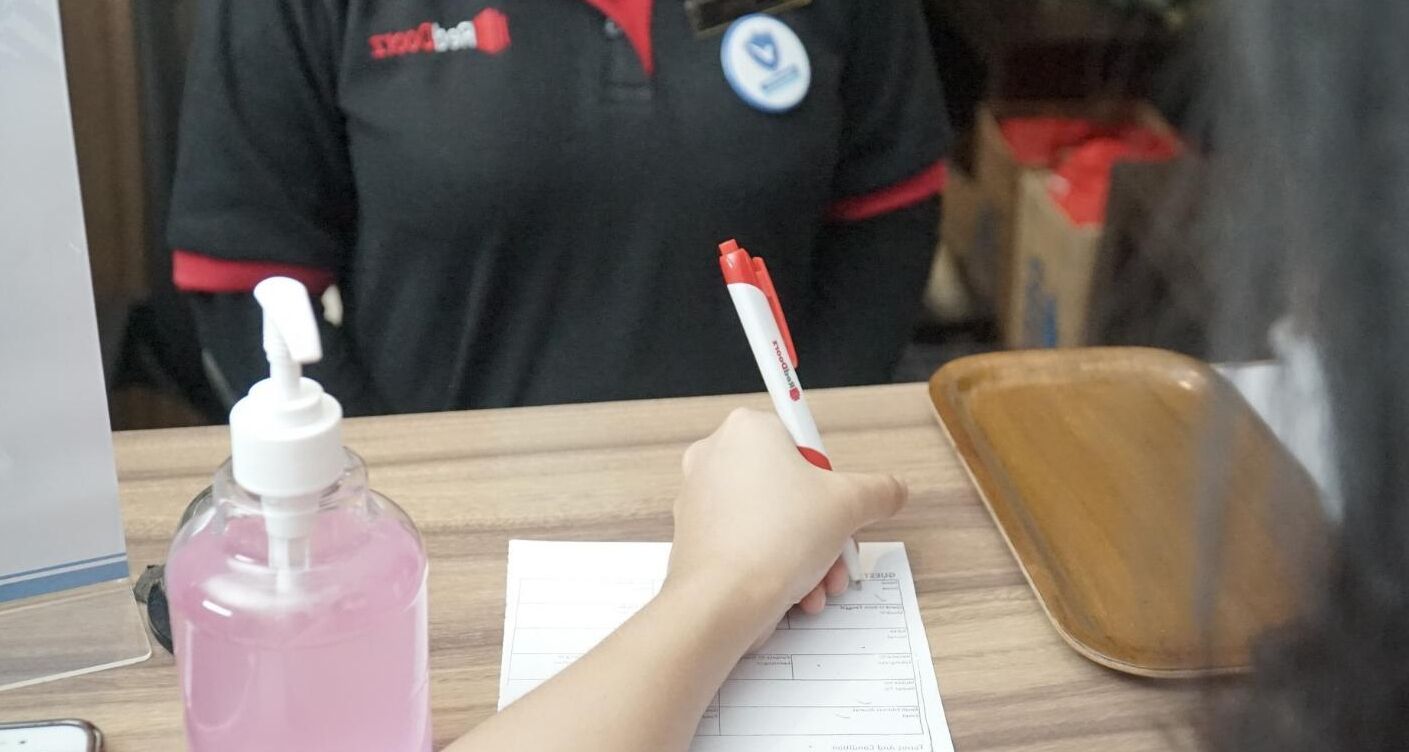 A hand holding a pen, writing on a paper, with a pink bottle and a brown tray on a wooden table.