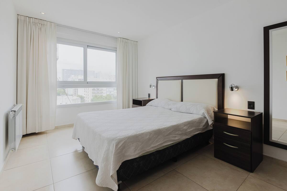 A bedroom with a large bed, a nightstand, a dresser, a mirror, and a window with curtains.