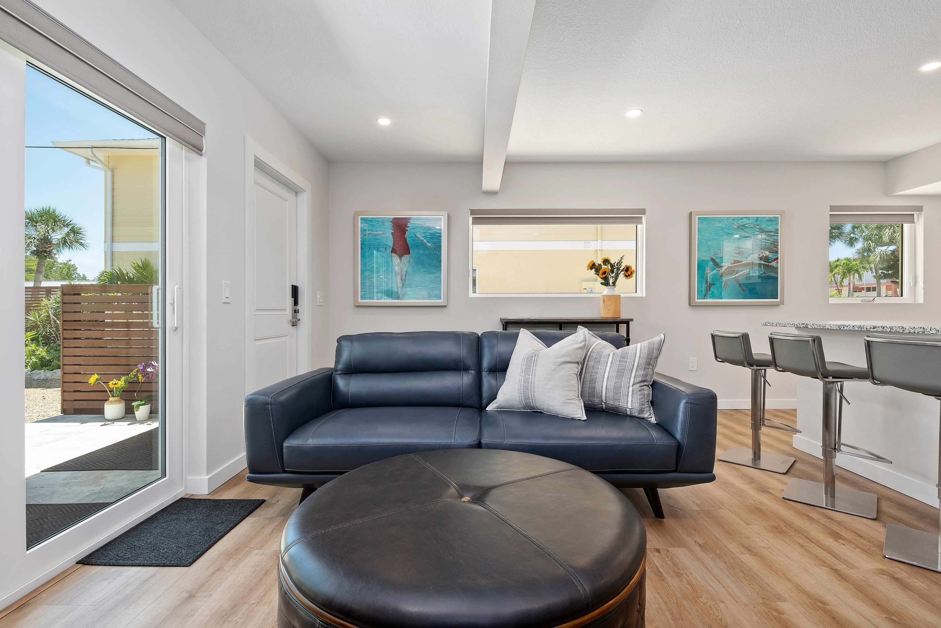 Una sala de estar moderna con un sofá de cuero azul oscuro, un otoman redondo y un comedor con taburetes de bar.