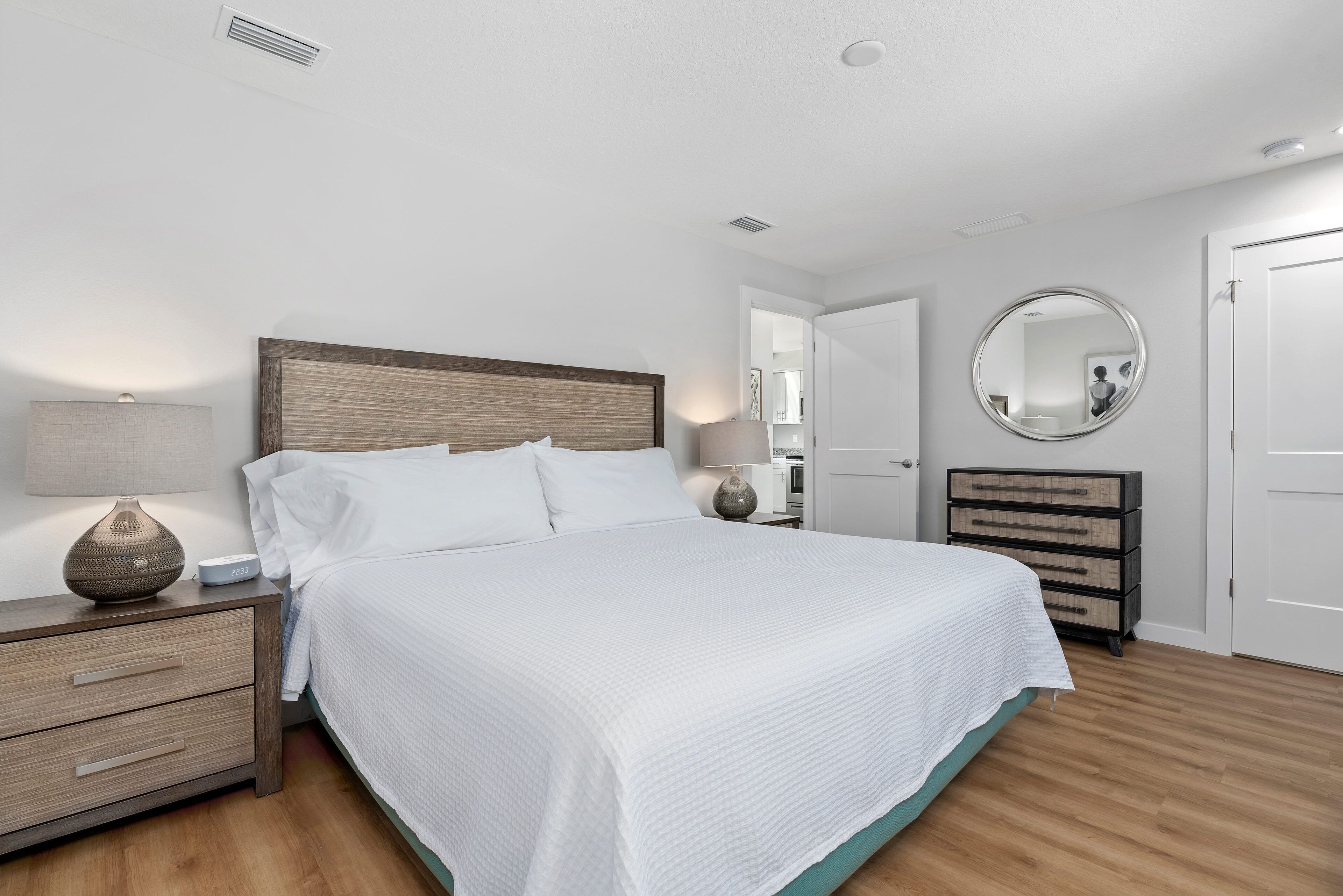 A bedroom with a large bed, a bedside table with a lamp, a dresser, and a mirror on the wall.