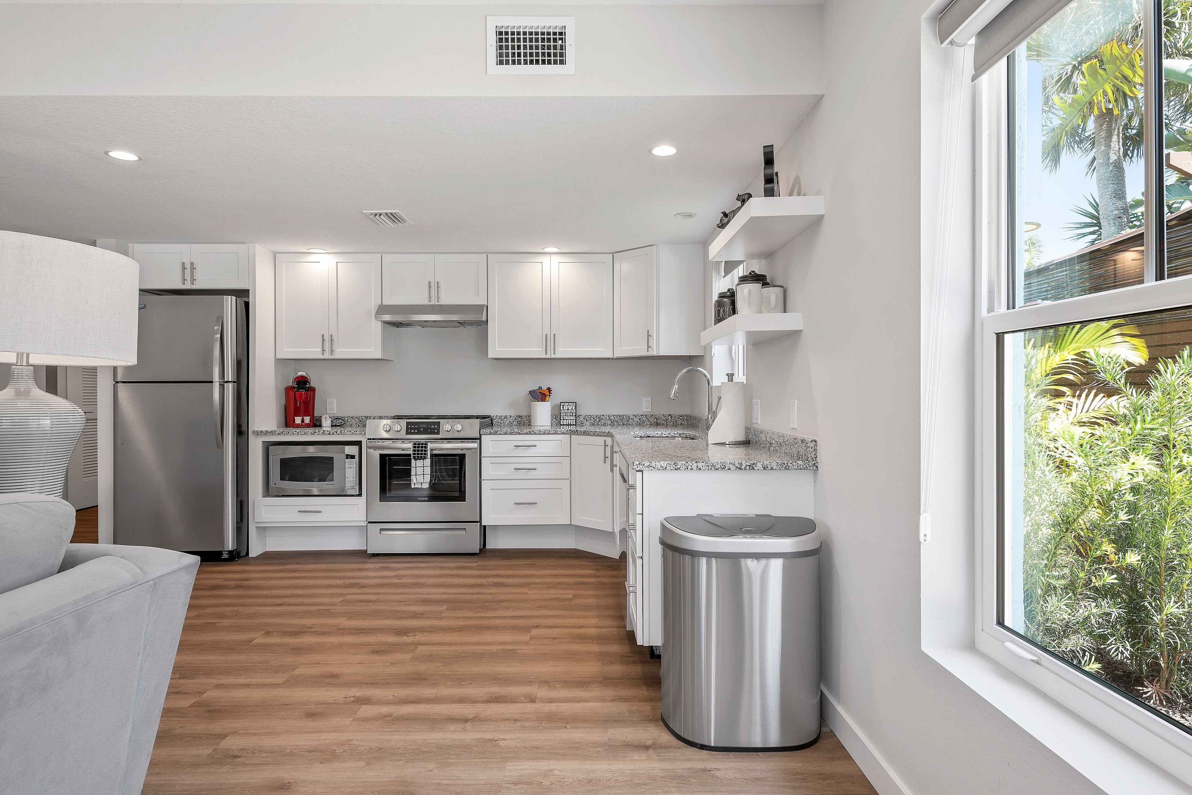 A modern kitchen with white cabinets, stainless steel appliances, and a window with a view of greenery.