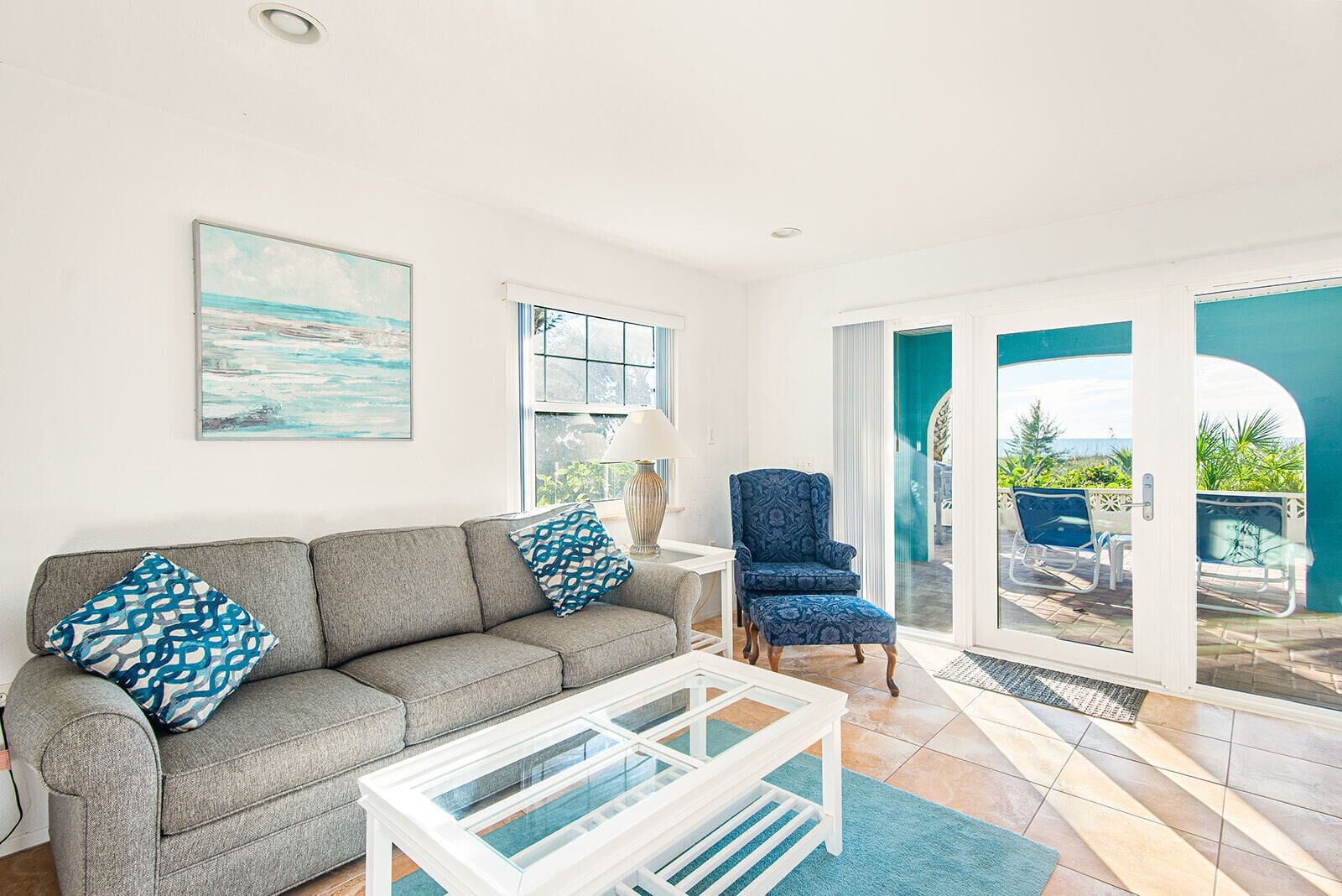Un salon comprenant un canapé, une table basse, un fauteuil et une vue sur un espace extérieur aménagé avec des chaises, accessible par une porte vitrée.