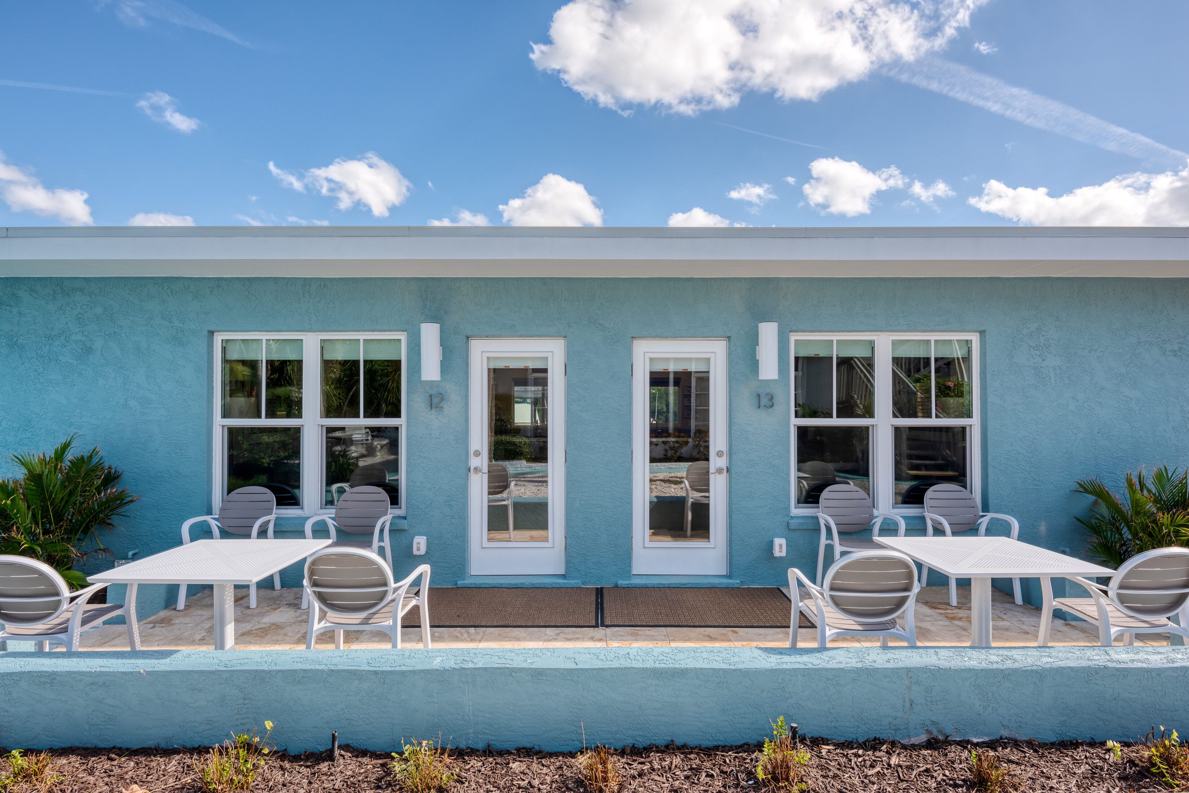 Un bâtiment à la façade bleu clair, doté de deux grandes fenêtres et d’un patio avec des tables et des chaises blanches.