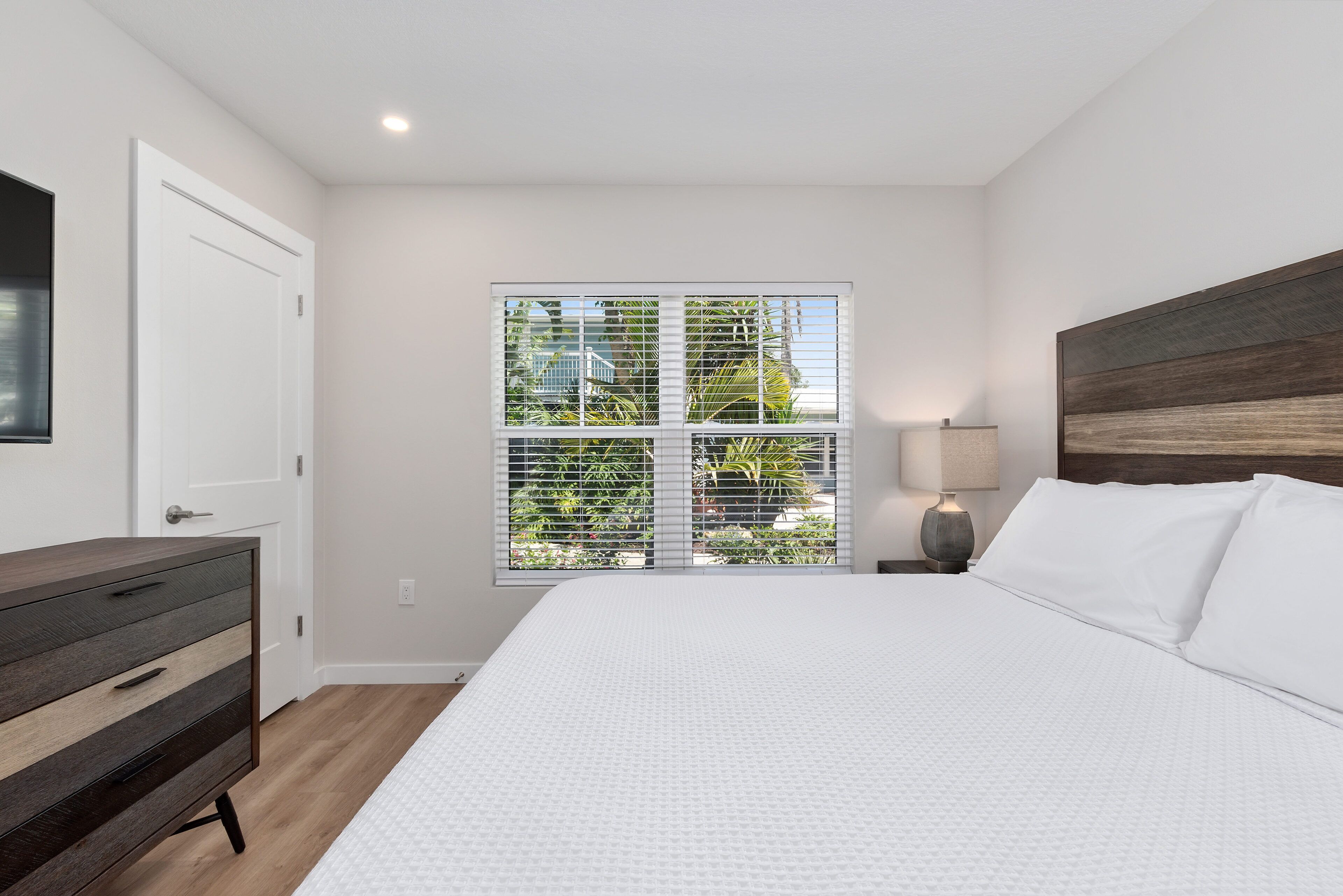 Un dormitorio con una cama grande, un aparador, una ventana con persianas y una mesita de noche con lámpara.