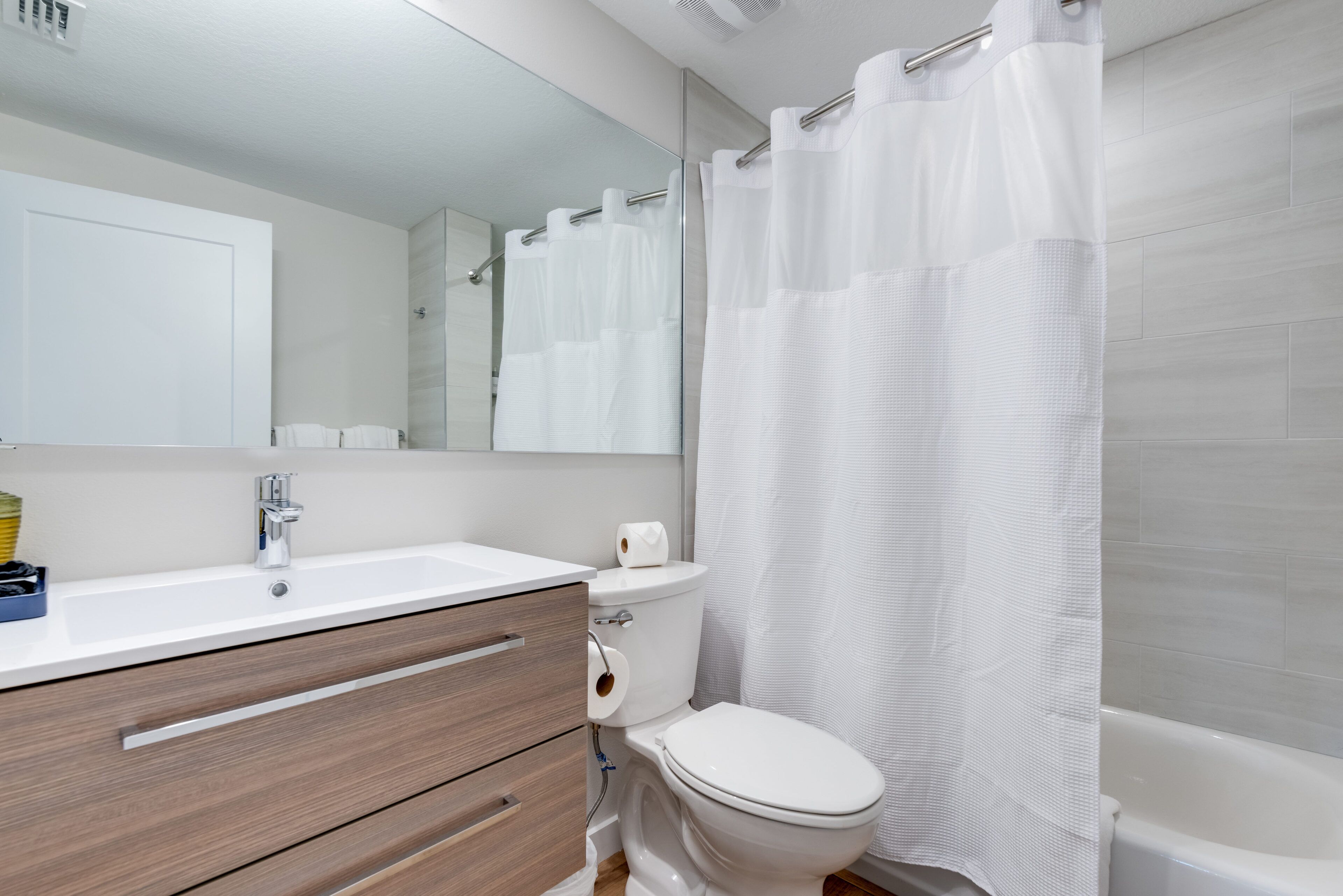 A modern bathroom with a white sink, wooden drawers, a toilet, and a shower curtain.