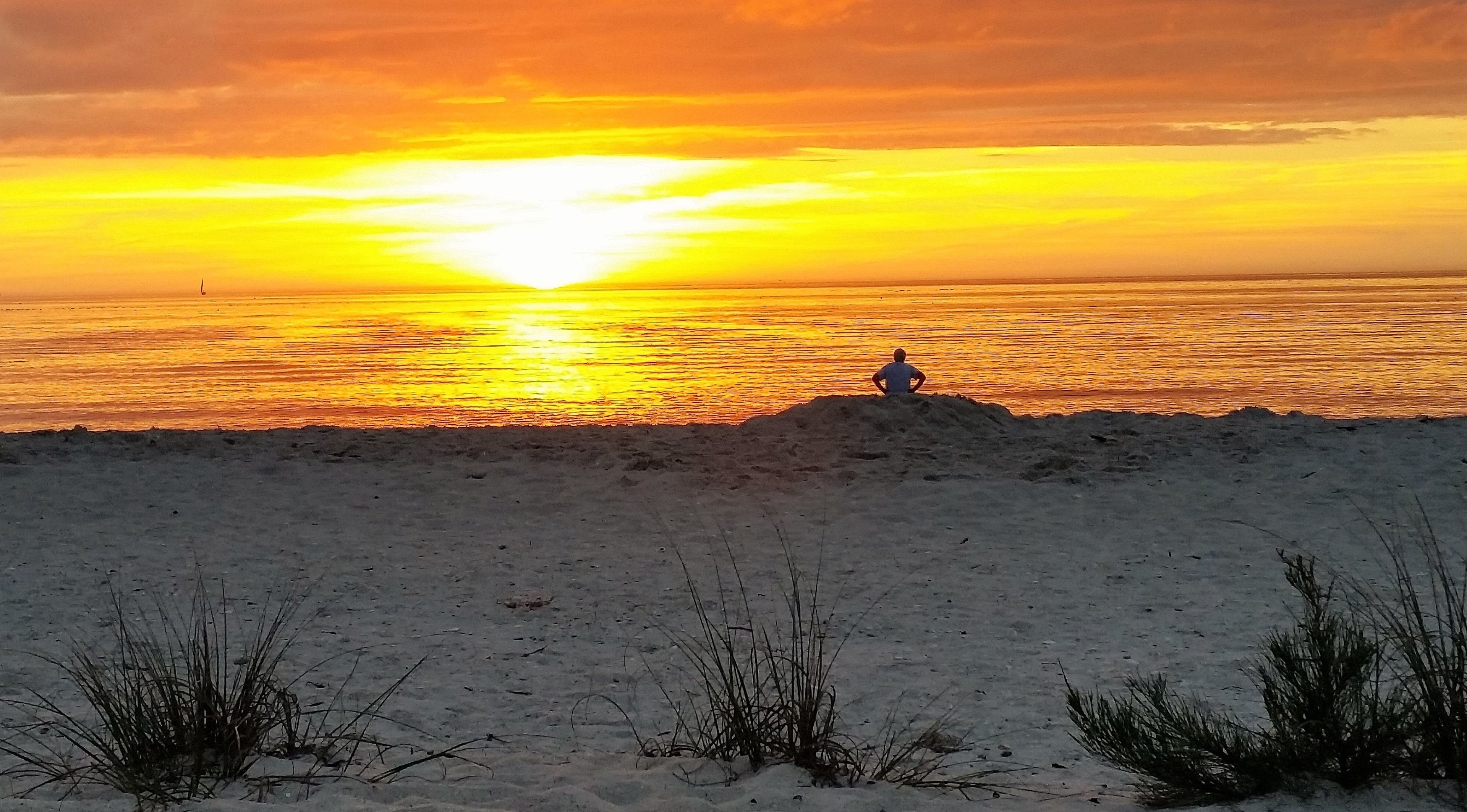 Una playa con una persona sentada en un montículo, contemplando el océano al atardecer.