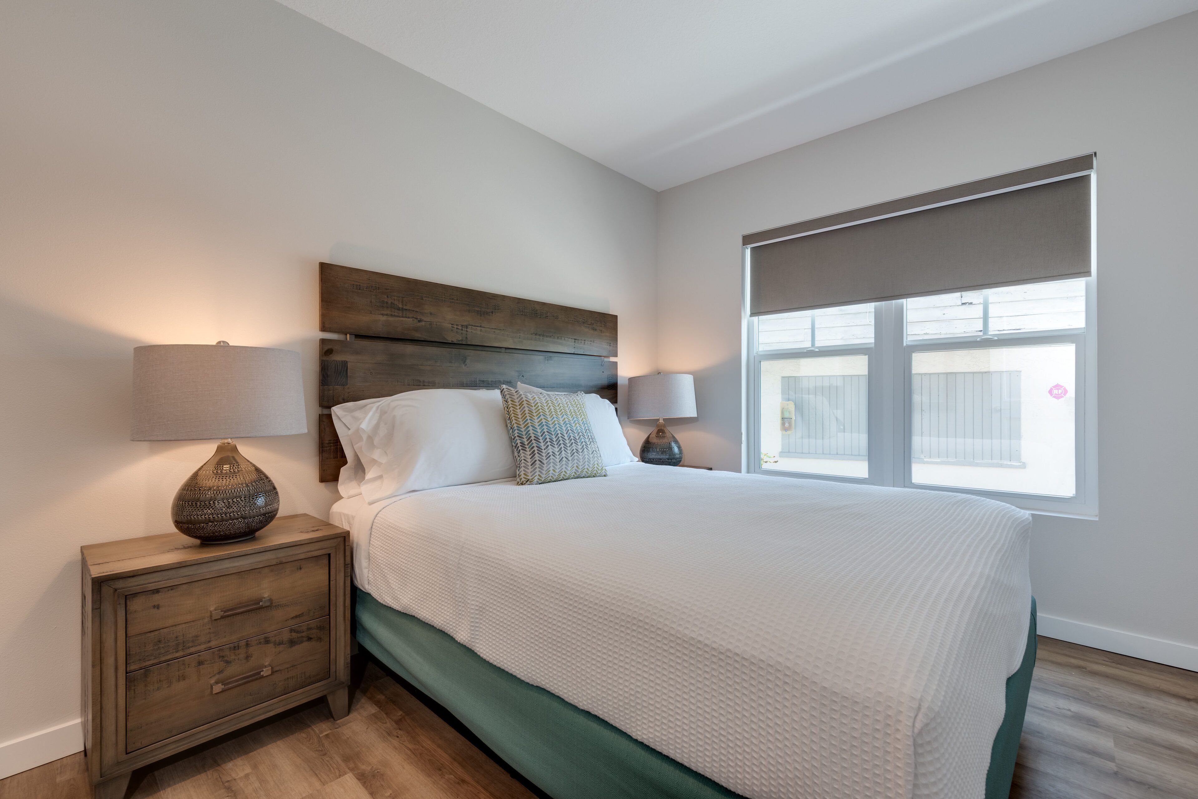 Une chambre à coucher avec une tête de lit en bois, un couvre-lit vert, deux lampes de chevet et une fenêtre avec un store.