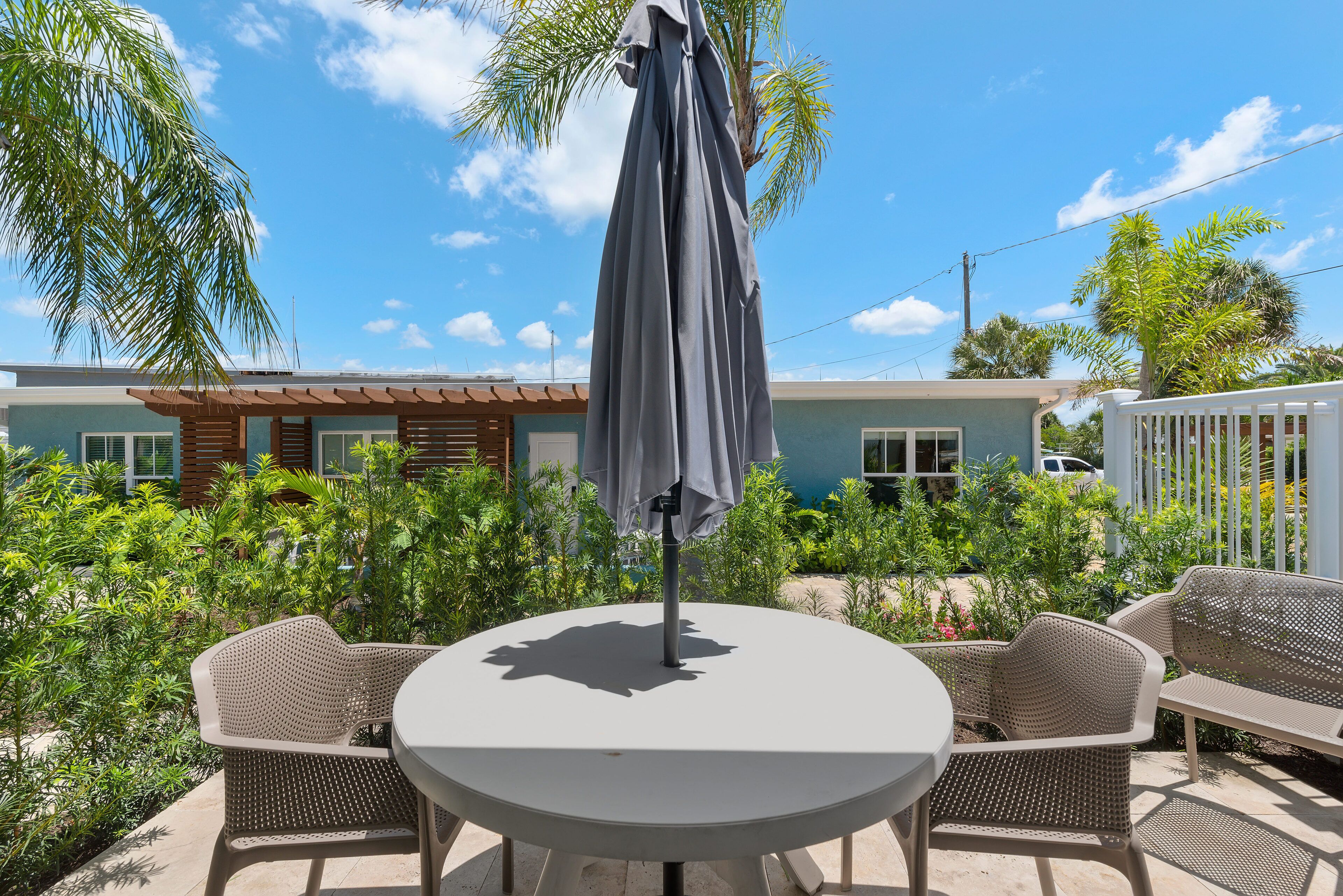 A round table with a grey umbrella, surrounded by wicker chairs, set on a patio with lush greenery and a building in the background.