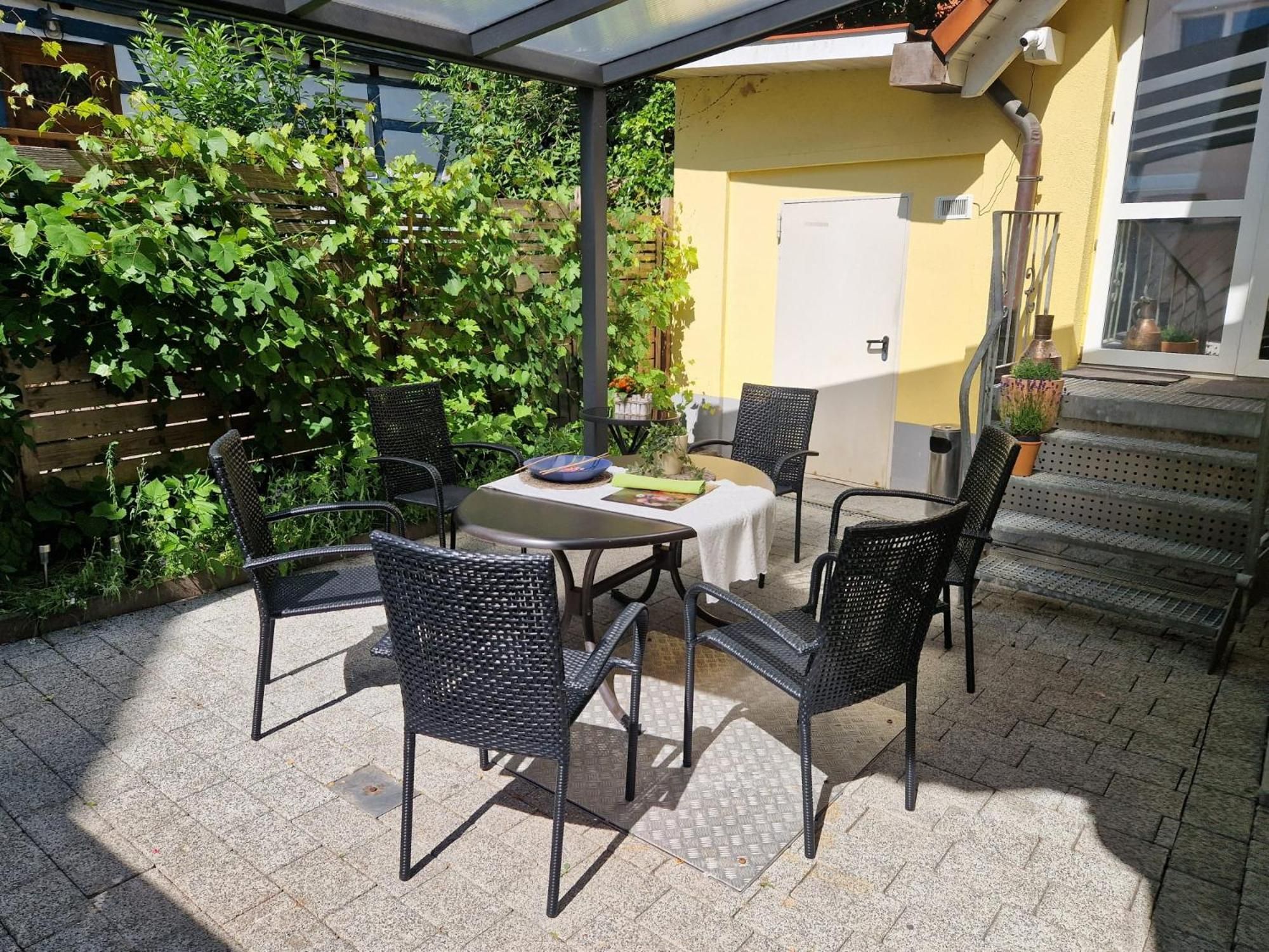 Eine Außenpatio mit Tisch und Stühlen, umgeben von Grünpflanzen und einem gelben Gebäude.