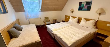 A hotel room with a large bed, a smaller bed, a desk, a chair, a radiator, and a window with blinds.
