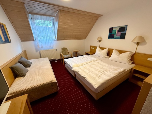 A hotel room with a large bed, a smaller bed, a desk, a chair, a radiator, and a window with blinds.