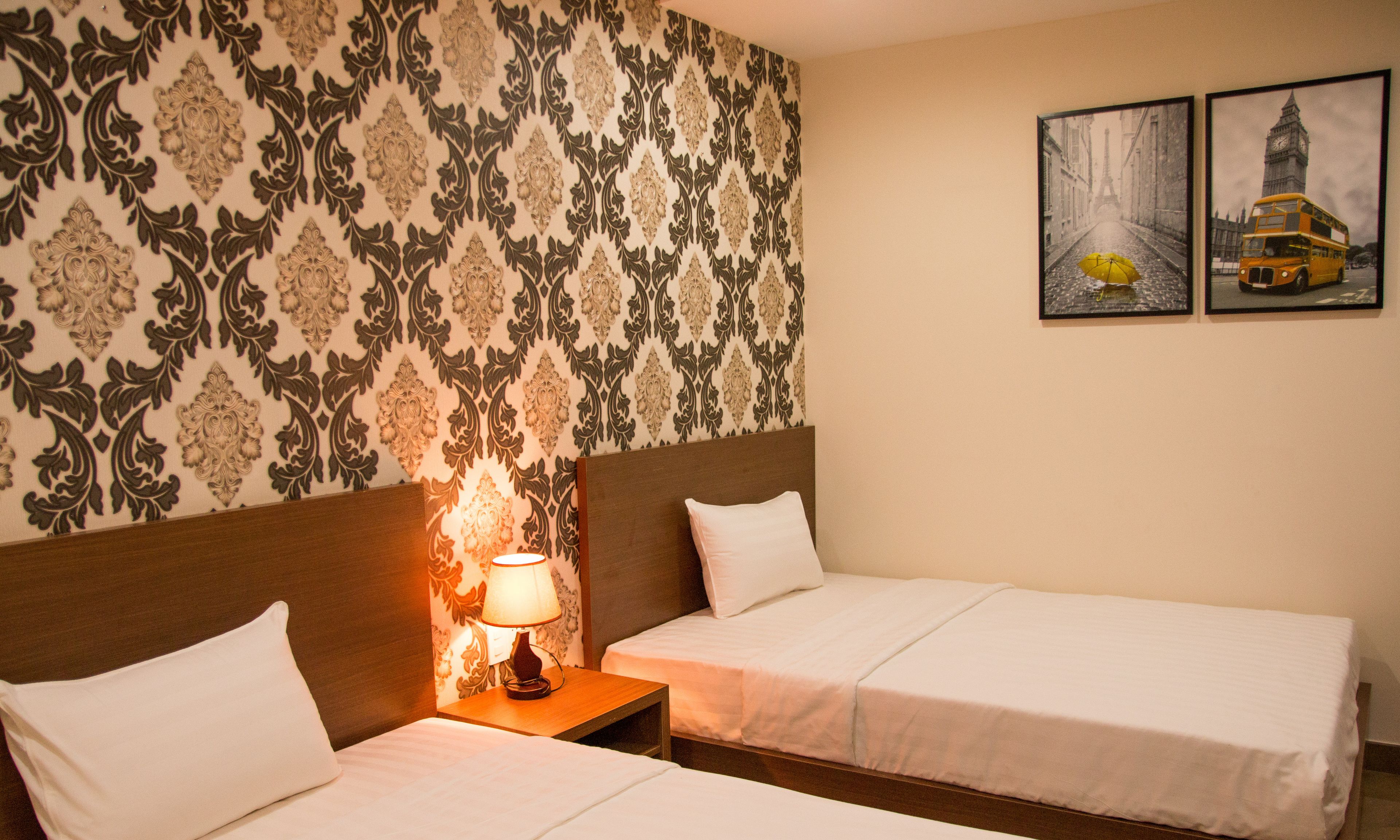 A hotel room with two single beds, a nightstand with a lamp, patterned wallpaper, and two framed pictures on the wall.