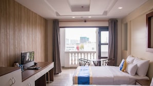 A hotel room with a bed, a desk with a TV, a balcony with a view of the city, and a dining area with chairs. - C'Lavie Hotel - Saigon Airport Hotel (Ho Chi Minh City)