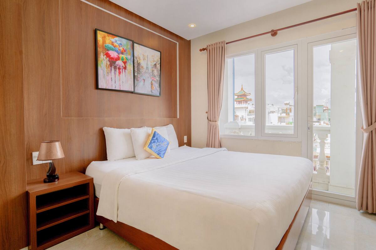 Ein Hotelzimmer mit einem großen Bett, einem Nachttisch, einer Lampe, einem Gemälde und Blick auf ein traditionelles Gebäude durch das Fenster.