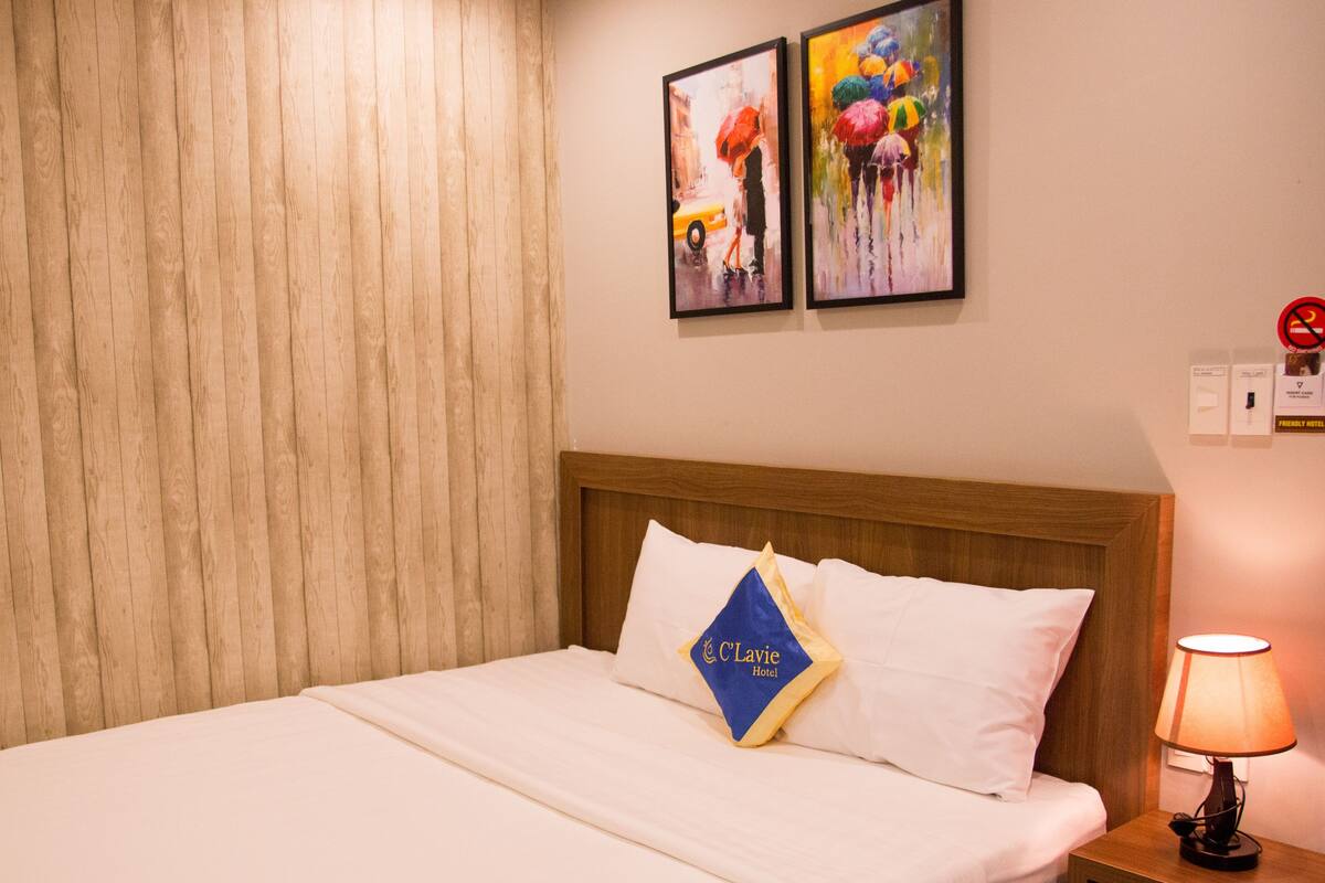 Ein Hotelzimmer mit einem Holzkopfteil, einem Bett mit weißen Leinentüchern, einem Kissen mit einem Logo, einem Nachttisch mit einer Lampe und zwei gerahmten Kunstwerken an der Wand.