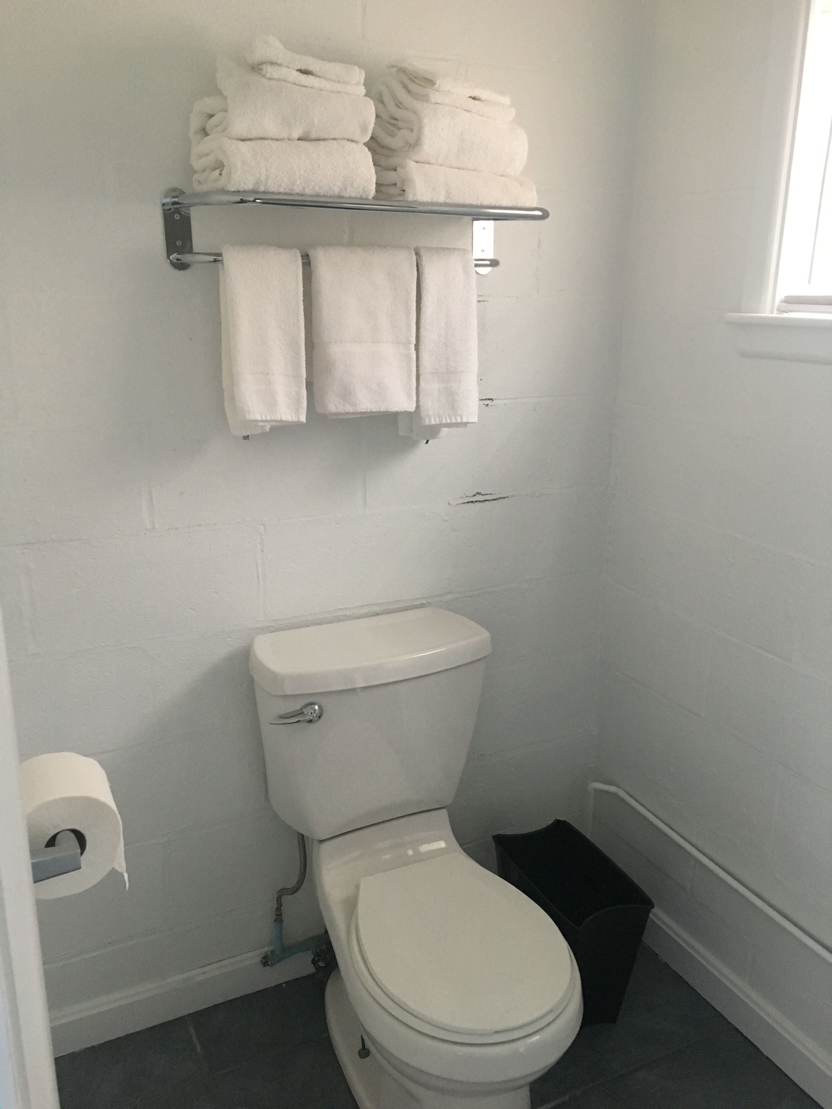 Ein Badezimmer mit Toilette, Ablagefläche mit gefalteten Handtüchern, einer Toilettenpapierrolle und einer schwarzen Mülleimer.