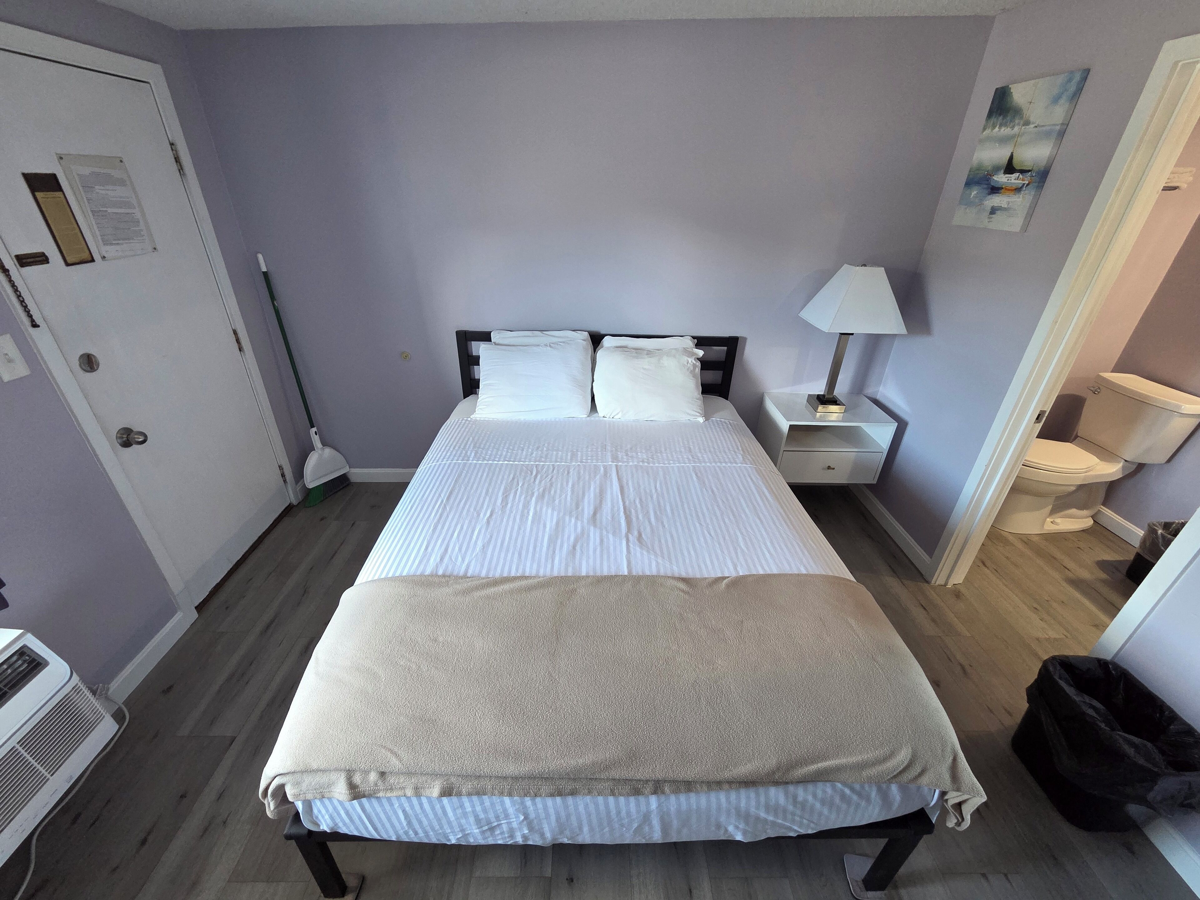 Una cama bien hecha con sábanas blancas, una mesita de noche con lámpara y un cuadro en la pared.