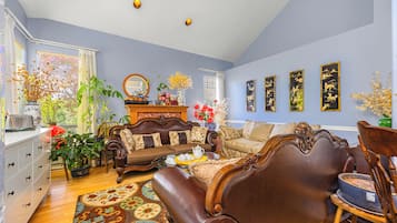 A living room with a brown leather sofa, a wooden coffee table, and a dining table with chairs. There are potted plants and a vase with flowers on the coffee table.