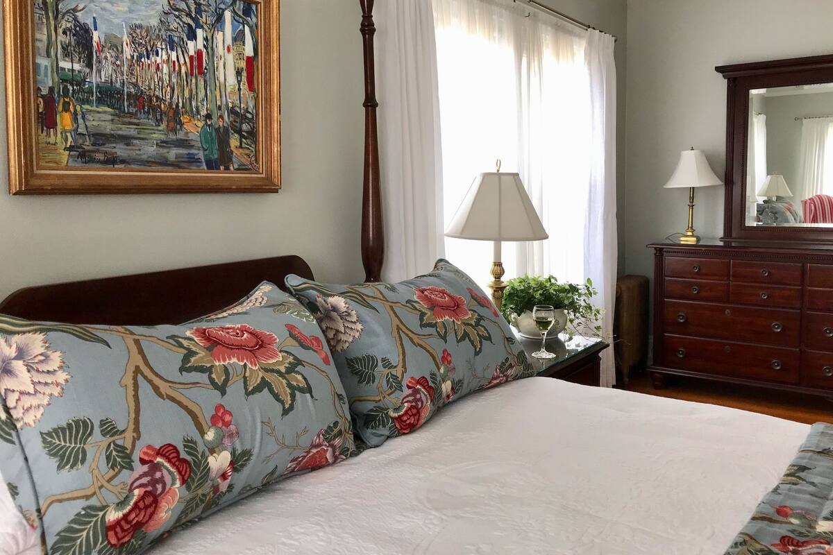 Ein ordentlich bezogenes Bett mit floralen Kissen, eine Stehlampe, eine Kommode mit Spiegel und ein Gemälde an der Wand.