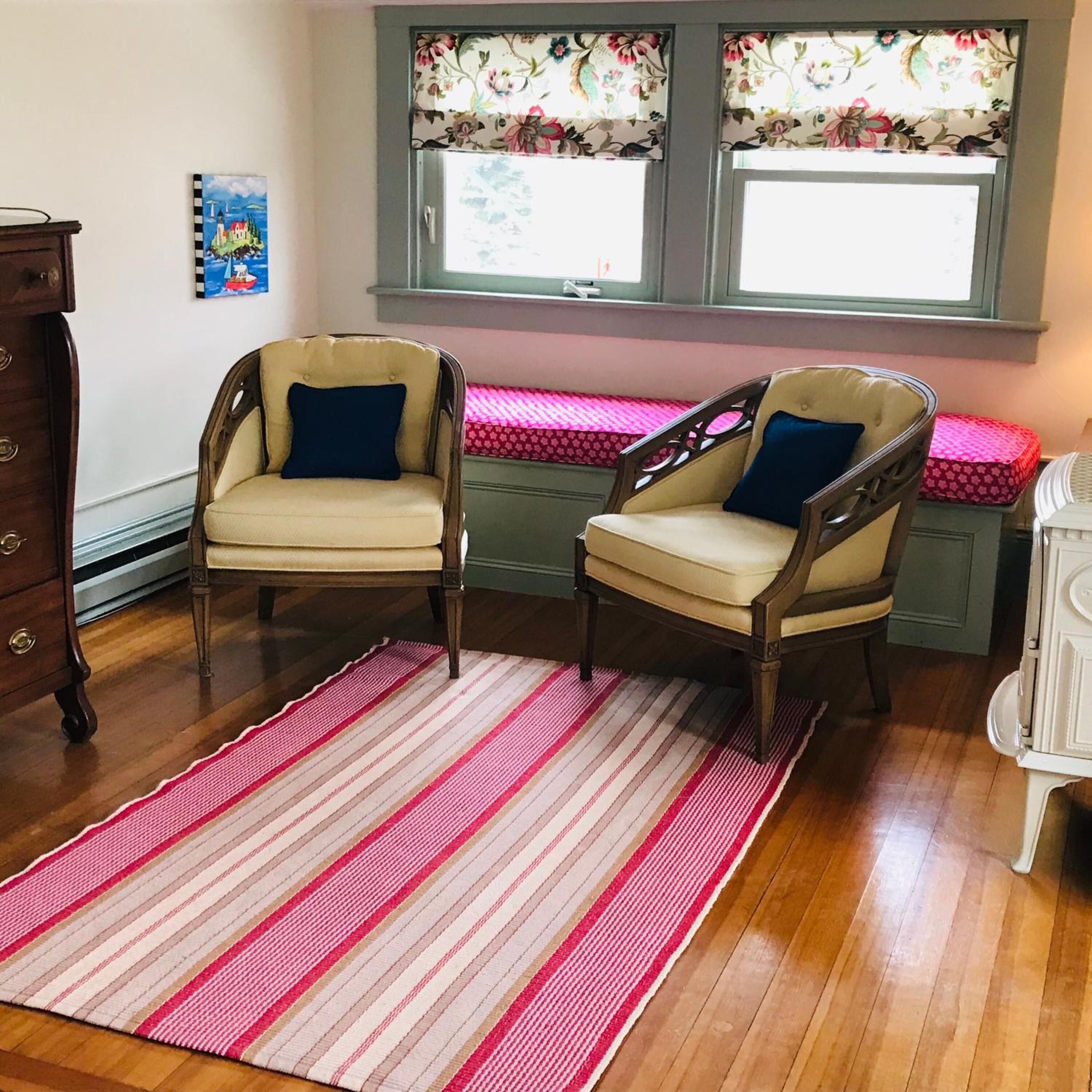 Quarto com duas cadeiras de madeira com almofadas, um tapete listrado, uma cômoda e uma janela com cortinas florais.