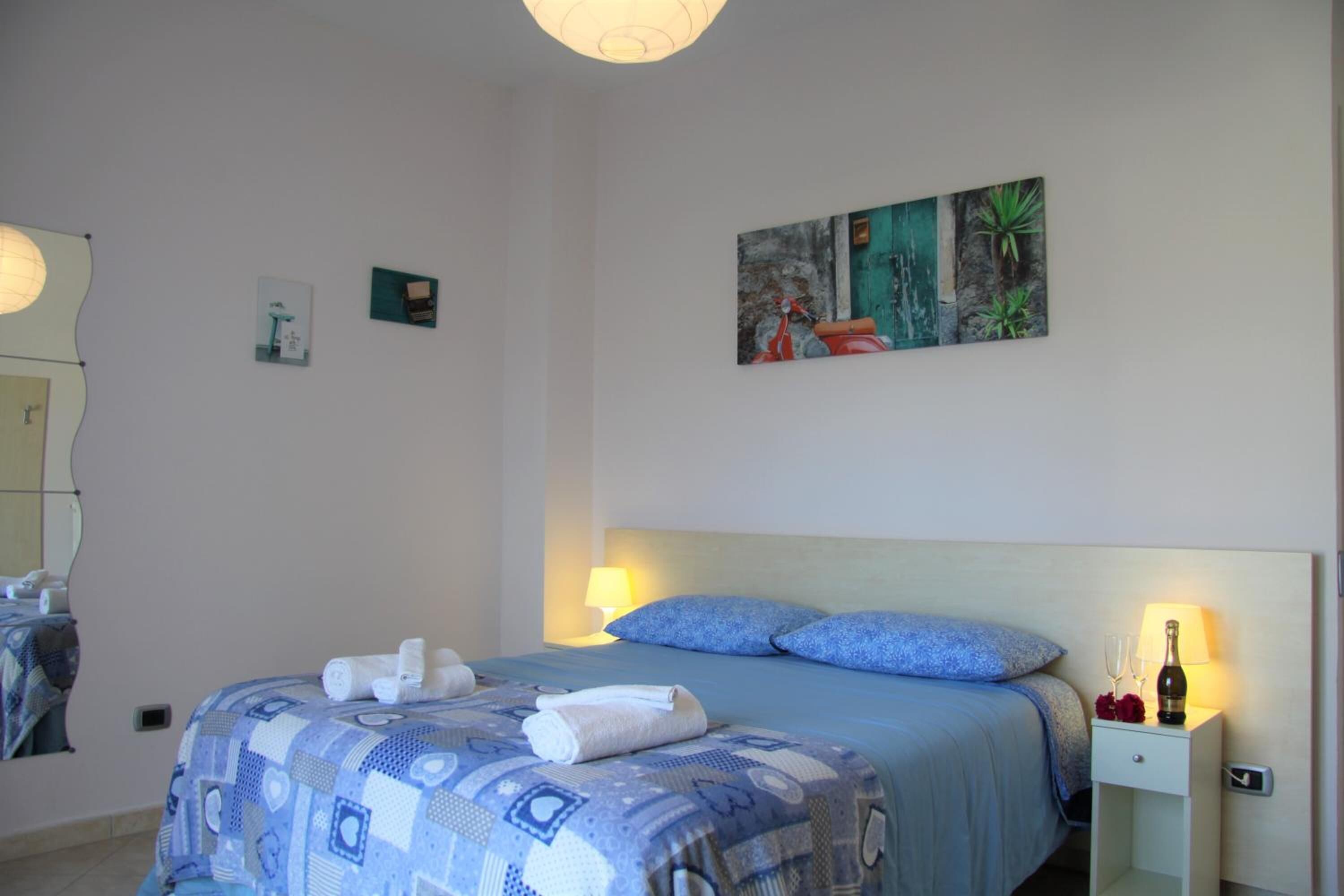 A neatly made bed with blue bedding and white towels, a bedside table with a bottle and glasses, and a wall with a painting.