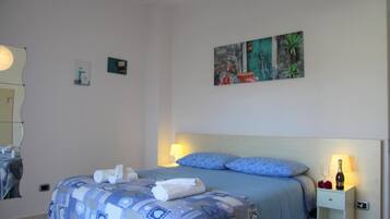 A neatly made bed with blue bedding and white towels, a bedside table with a bottle and glasses, and a wall with a painting.