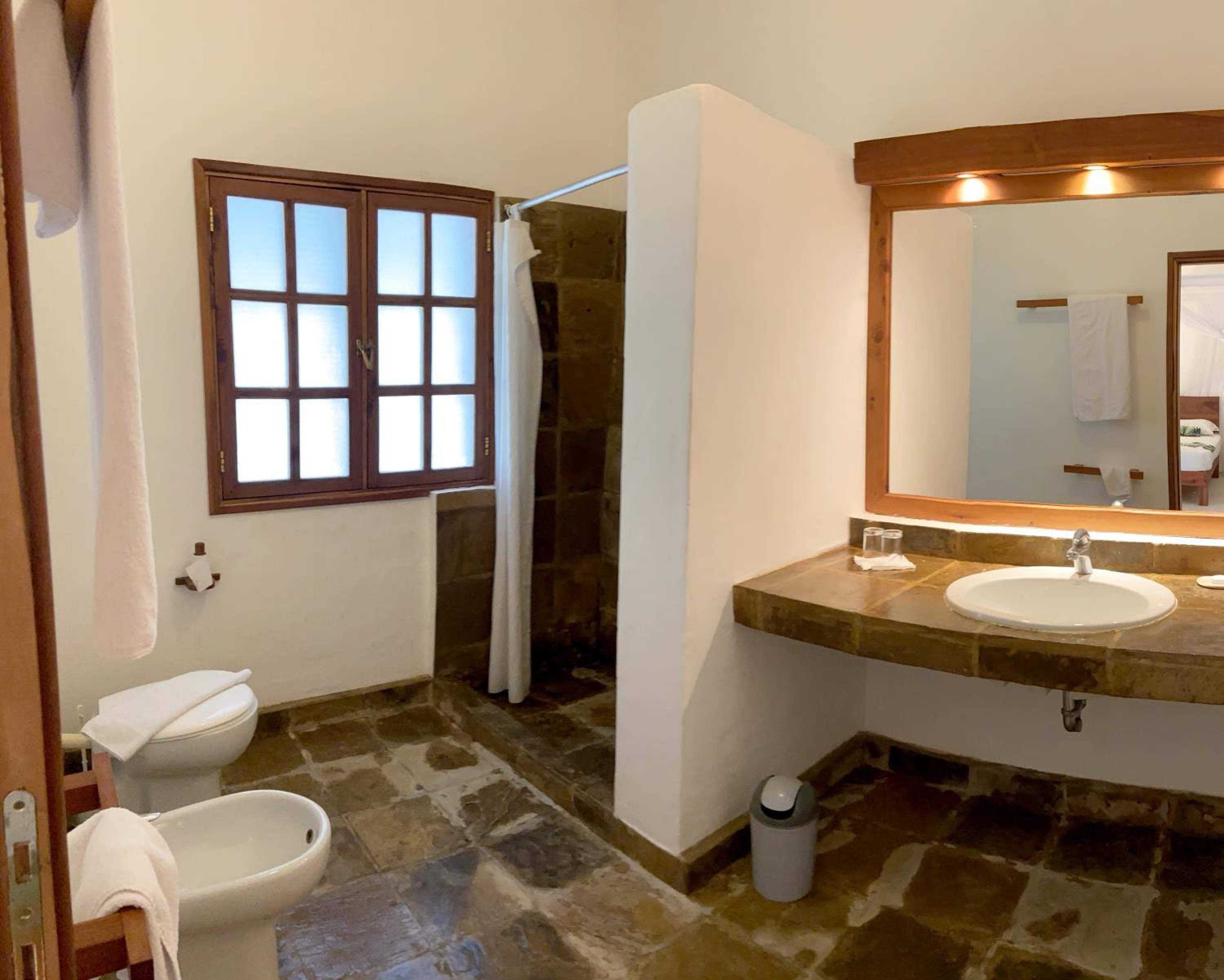 A bathroom with a stone floor, a white sink, a mirror, and a window with frosted glass.