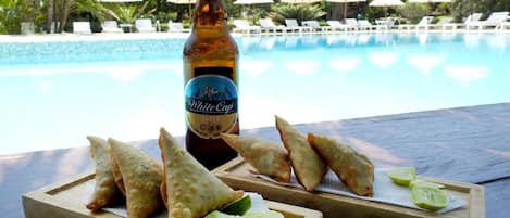 A bottle of White Cap Lager and samosas on a wooden board with lime wedges, overlooking a pool with lounge chairs and umbrellas.