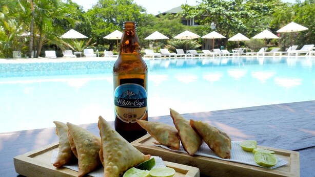 A bottle of White Cap Lager and samosas on a wooden board with lime wedges, overlooking a pool with lounge chairs and umbrellas.