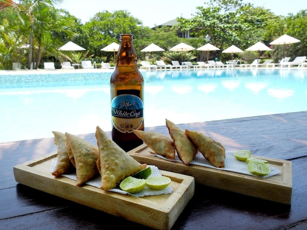 A bottle of White Cap Lager and samosas on a wooden board with lime wedges, overlooking a pool with lounge chairs and umbrellas.