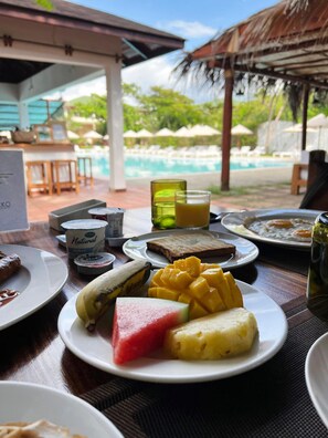 Impostazione colazione con piatti di cibo, tra cui pane tostato, frutta e una bevanda, in un'area esterna soleggiata con piscina e ombrelloni sullo sfondo.