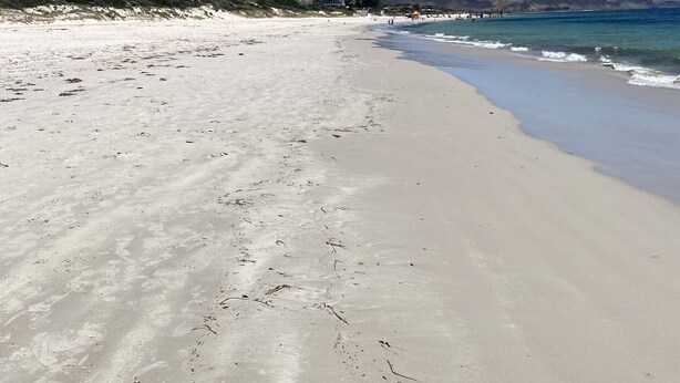 A wide sandy beach with gentle waves, a clear blue sky, and distant hills.