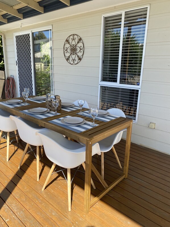 A wooden deck with a dining table set for four, white chairs, and a decorative wall piece.