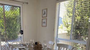 A dining area with a wooden table set for six, white chairs, and a view of greenery through the window.