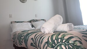 Een bed met een bedspreider met een tropisch bladmotief, witte kussens en twee opgevouwen handdoeken.