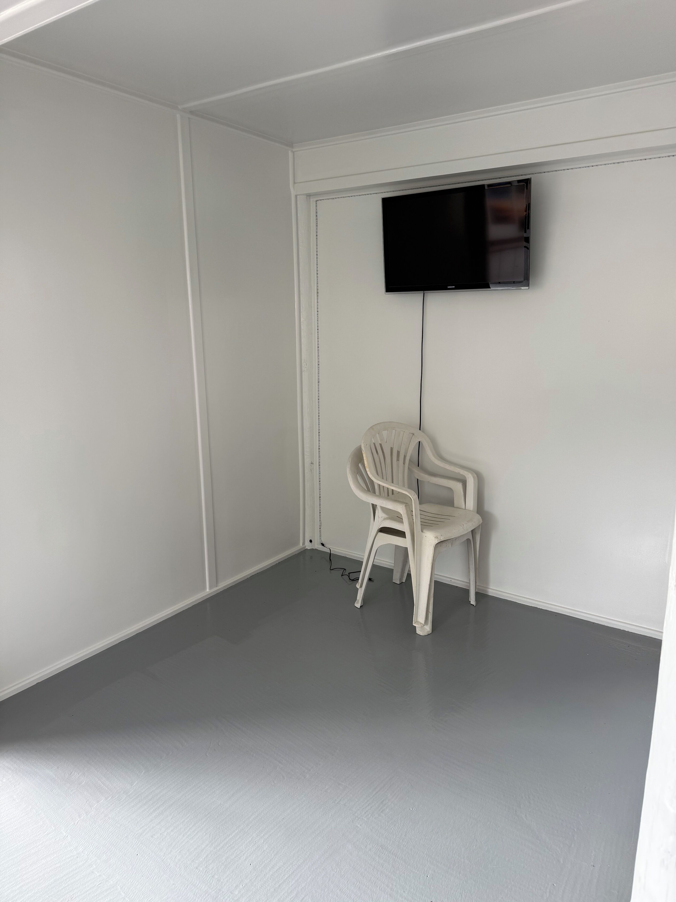 A room with white walls, a grey floor, a white plastic chair, and a mounted television.
