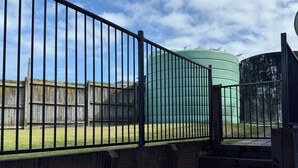 A set of outdoor stairs leading to a fenced area with a grassy lawn and a large green water tank in the background. - Tokerau Beach Paradise (Tokerau Beach)