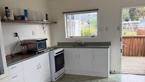 A compact kitchen with white cabinets, a built-in oven, a microwave, and a small countertop. There is a window above the sink, and a door leading to a backyard. - Tokerau Beach Paradise (Tokerau Beach)
