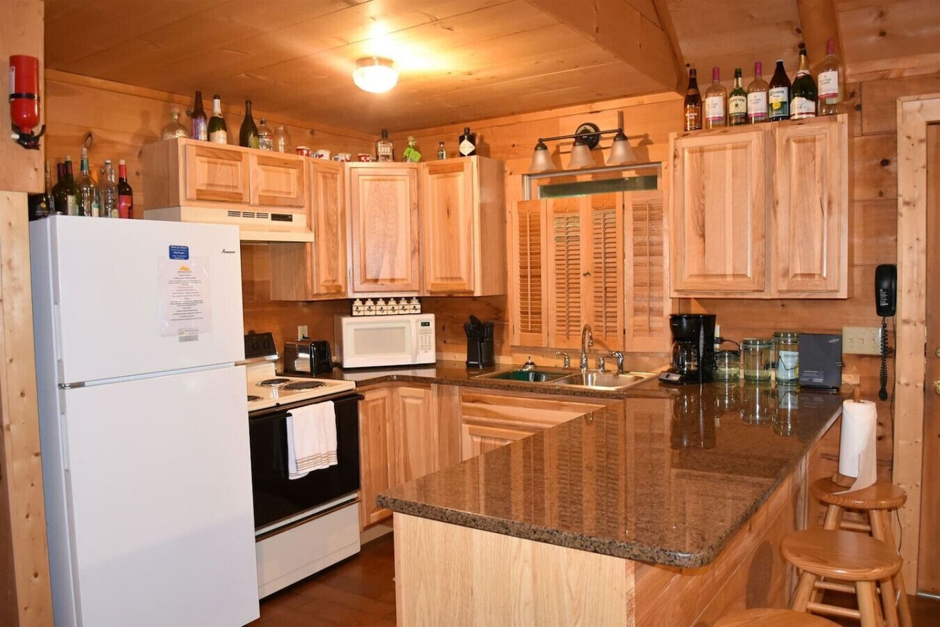 Una cocina con armarios de madera, un refrigerador blanco, un microondas, una estufa y un lavaplatos. Hay taburetes y una encimera con botellas.