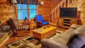 A cozy cabin interior with a wooden interior, a sofa, a blue chair, a wooden coffee table, and a television on a stand. - Trail`s End-Hot tub-Wi-Fi,-pets ok-Pool Table-Fireplace-Screened Porch-Activities (Morganton)