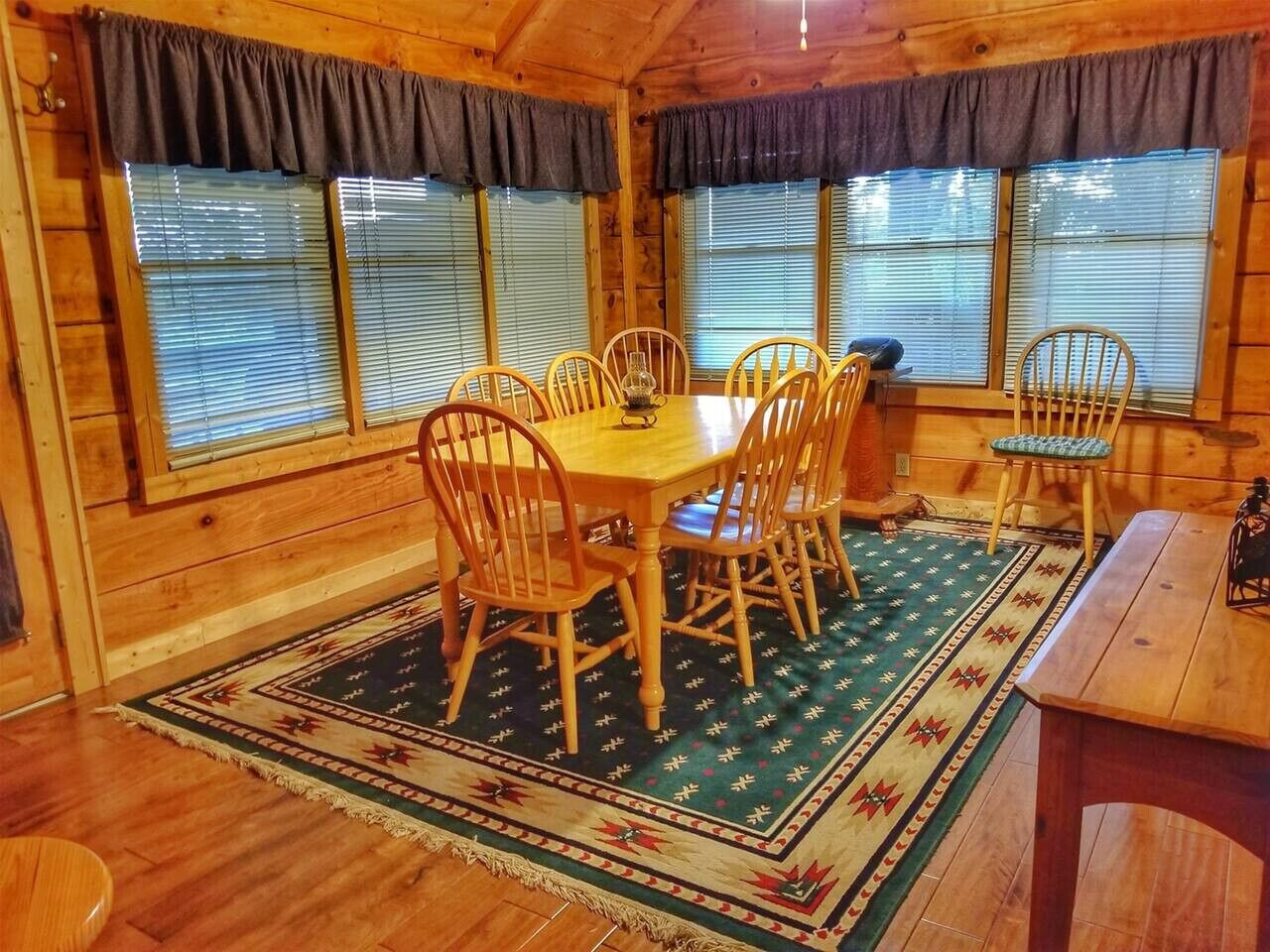 Een houten chalet van binnen, met een eettafel omringd door stoelen, een vloerkleed met dessin en grote ramen met zonwering.