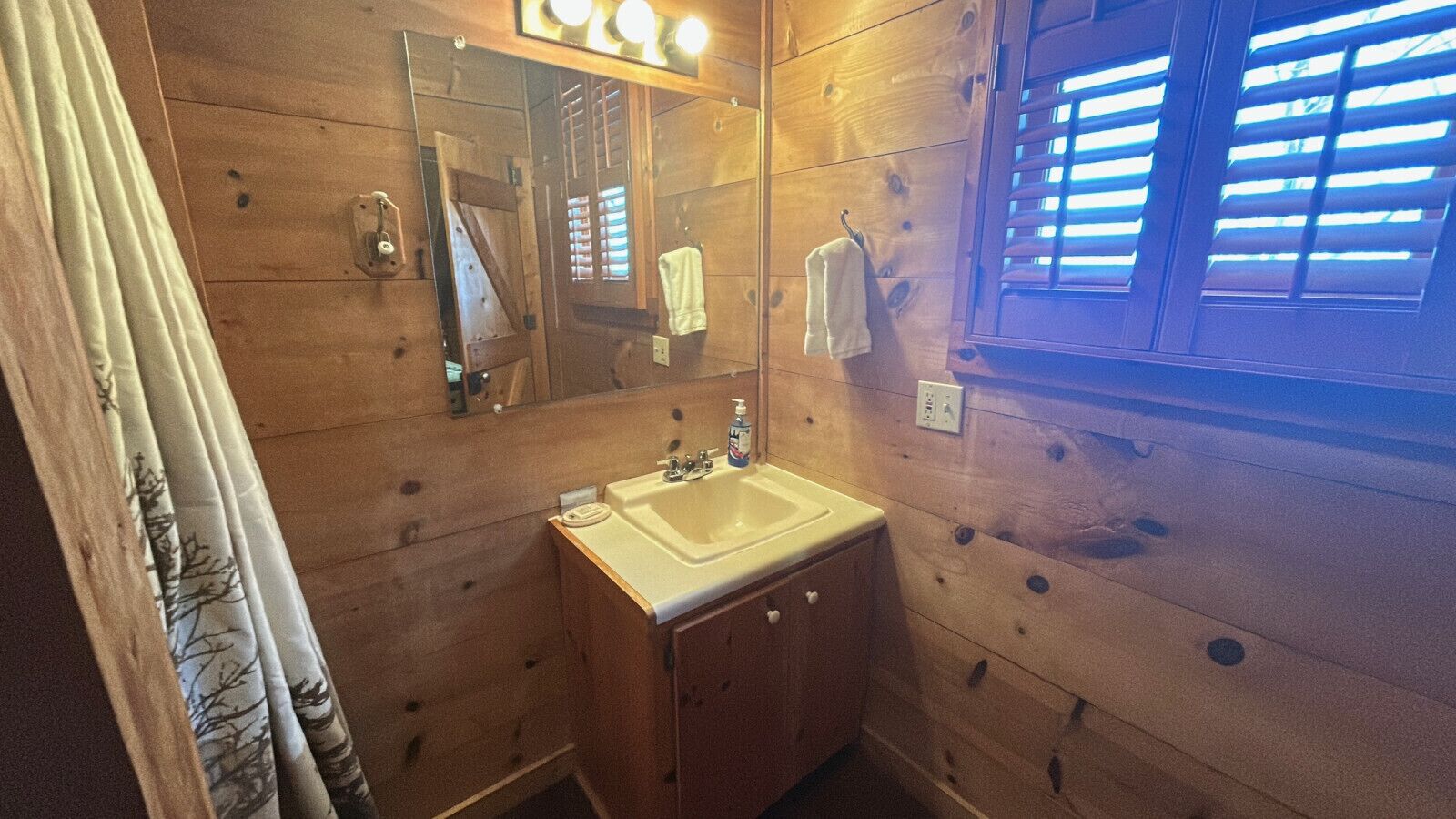 A wooden cabin bathroom with a sink, mirror, and window with blinds.