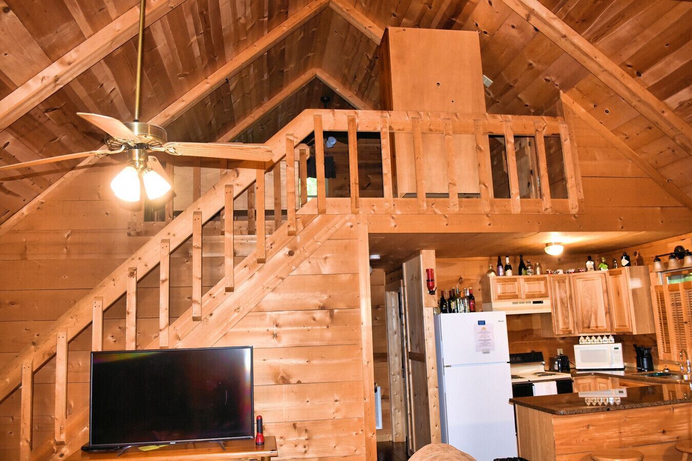 Een houten chalet van binnen met een trap, een televisie, een keukenhoek en een plafondventilator.