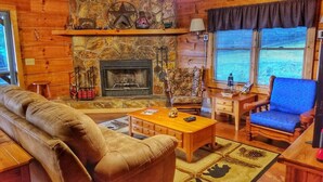 A cozy cabin interior with a stone fireplace, wooden furniture, and a large window with blinds. - Trail`s End-Hot tub-Wi-Fi,-pets ok-Pool Table-Fireplace-Screened Porch-Activities (Morganton)
