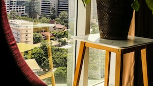 Una ventana con vista a un paisaje urbano, una planta en maceta sobre una mesita y una silla roja en primer plano.
