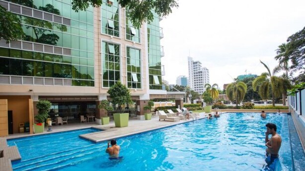 Zona de piscina con un edificio moderno al fondo, sillones de descanso y cielo despejado.