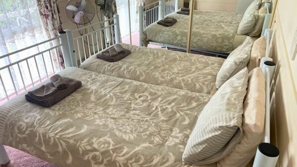 Une chambre double avec deux lits, chacun recouvert d’une courtepointe à motifs floraux, avec des oreillers et une serviette pliée. La chambre est équipée d’une fenêtre avec des rideaux, d’un ventilateur et d’un radiateur.